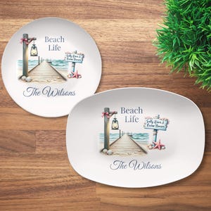 May include: Two white ceramic plates with a beach theme. One is round, the other oval. Each features a pier illustration with the words "Beach Life" and "The Wilsons". A sign reads "Salty Kisses & Ocean Breezes".