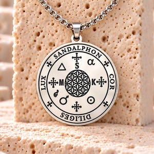 May include: A silver-toned circular pendant necklace featuring an intricate black engraved design. The centre displays a flower of life pattern surrounded by symbols, letters, and the text SANDALPHON, LUX, COR, and DILIGES on a polished metal surface.