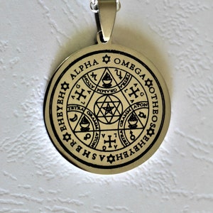 May include: A silver pendant necklace with a round, silver charm. The charm has a black inscription that reads "ALPHA OMEGA OTHBOS EHHVBH", and features a star of David, a cross, and other symbols.