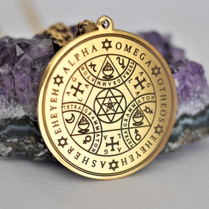 May include: A gold-colored pendant necklace with a circular charm featuring a complex design of symbols and text. The text includes "ALPHA", "OMEGA", "OTHEOS", and "EHEYEH", among others.