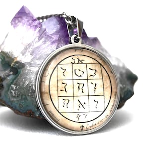 May include: A round silver-toned pendant necklace featuring a beige circular insert with a black grid containing various symbols and Hebrew-style characters, suspended in front of a large, jagged, translucent purple amethyst crystal cluster.