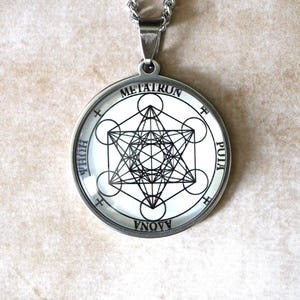 K&ouml;nnte beinhalten: Ein runder, silberfarbener Anh&auml;nger an einer Kette mit einem schwarz-wei&szlig;en geometrischen Metatron-W&uuml;rfel-Design. Der &auml;u&szlig;ere Rand zeigt den Text METATRUN, PUIA, AAONA und HUHA in schwarzen Gro&szlig;buchstaben auf wei&szlig;em Hintergrund.