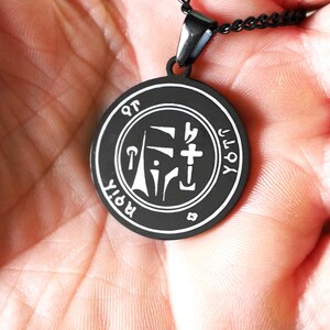 May include: Black pendant necklace with a silver sigil charm. The sigil is a geometric design with a cross and the words "ROIT" and "YOT" around the outside.