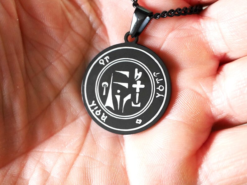 May include: Black pendant necklace with a silver sigil charm. The sigil is a geometric design with a cross and the words "ROIT" and "YOT" around the outside.
