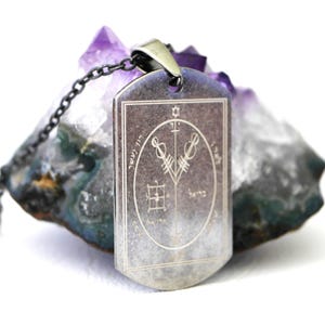 Può includere: Una collana con pendente rettangolare color argento con un design inciso dettagliato. Il pendente è appeso a una catena scura e presenta un emblema circolare centrale con simboli e testo. Lo sfondo include un cristallo viola e grigio.