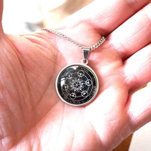 K&ouml;nnte beinhalten: Eine silberfarbene runde Anh&auml;ngerkette mit einem schwarz-wei&szlig;en geometrischen Metatron-W&uuml;rfel-Design unter einer Glaskuppel. Der Anh&auml;nger ist in eine schlichte Metallfassung eingefasst und h&auml;ngt an einer passenden silberfarbenen Gliederkette.