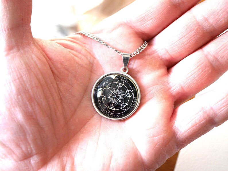 K&ouml;nnte beinhalten: Eine silberfarbene runde Anh&auml;ngerkette mit einem schwarz-wei&szlig;en geometrischen Metatron-W&uuml;rfel-Design unter einer Glaskuppel. Der Anh&auml;nger ist in eine schlichte Metallfassung eingefasst und h&auml;ngt an einer passenden silberfarbenen Gliederkette.