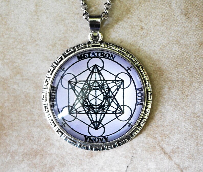 K&ouml;nnte beinhalten: Ein silberfarbener, runder Anh&auml;nger mit einem schwarzen, geometrischen Metatron-W&uuml;rfel-Design auf hellviolettem Hintergrund. Der Metallrahmen hat eine Bord&uuml;re mit M&auml;ander-Muster. Der Text METATRON steht oben auf dem runden Einsatz.