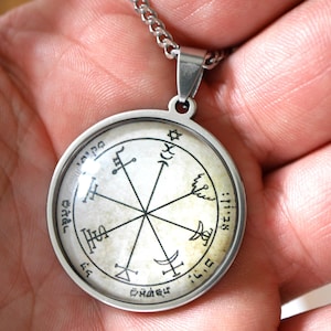 May include: A silver-colored pendant necklace featuring a circular charm with a detailed, intricate design. The charm has a central starburst pattern with symbols and text around the perimeter. The necklace is held by a person's hand.