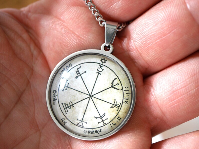 May include: A silver-colored pendant necklace featuring a circular charm with a detailed, intricate design. The charm has a central starburst pattern with symbols and text around the perimeter. The necklace is held by a person's hand.