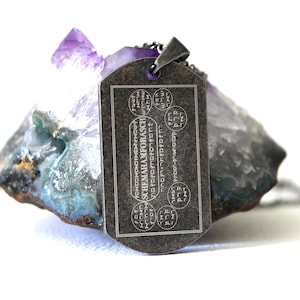 Pode incluir: Um pingente retangular cinza escuro com o texto "SCHEMA HAMFORASCH" e outros símbolos é exibido. O pingente pende de uma corrente prateada. O pingente repousa sobre uma pedra áspera multicolorida com um cristal roxo em cima.
