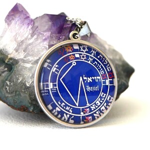 May include: A round, silver-toned pendant with a blue center featuring white Hebrew text and symbols. The pendant is suspended from a silver chain and rests on a purple and gray crystal. The word "Haziel" is visible.