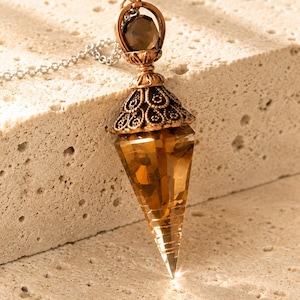 Peut inclure: Un pendentif en cristal facetté de couleur ambre, de forme pointue, suspendu à une chaîne argentée. Le cristal est surmonté d'un capuchon en métal cuivré orné de motifs en filigrane complexes et d'une petite perle facettée foncée.