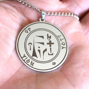 May include: Silver pendant necklace with a round, engraved charm featuring a symbol and the text "YHVH" and "MORYA".