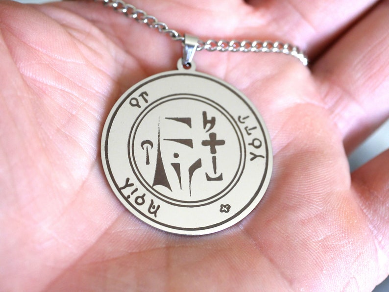 May include: Silver pendant necklace with a round, engraved charm featuring a symbol and the text "YHVH" and "MORYA".