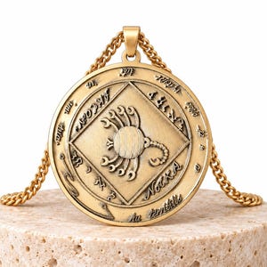 Könnte beinhalten: Eine goldfarbene, runde Anhängerkette mit einem zentralen, geprägten Skorpion in einer Rautenform. Der äußere Rand ist mit eingravierten Schriftzügen, darunter das Wort Nocka, und verschiedenen Symbolen verziert.