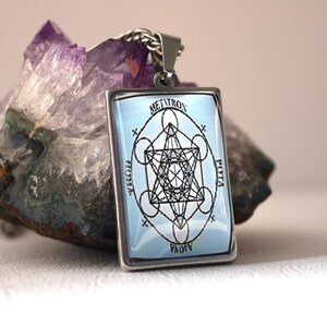 K&ouml;nnte beinhalten: Ein rechteckiger silberfarbener Anh&auml;nger mit hellblauem Hintergrund, einem schwarzen geometrischen Metatron-W&uuml;rfel-Design und dem Text METATRON, POTA und AJOVA um das zentrale Symbol, befestigt an einer silberfarbenen Metallkette.