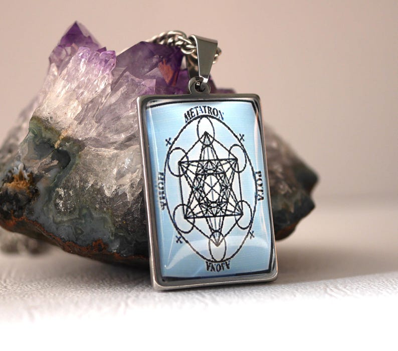 K&ouml;nnte beinhalten: Ein rechteckiger silberfarbener Anh&auml;nger mit hellblauem Hintergrund, einem schwarzen geometrischen Metatron-W&uuml;rfel-Design und dem Text METATRON, POTA und AJOVA um das zentrale Symbol, befestigt an einer silberfarbenen Metallkette.