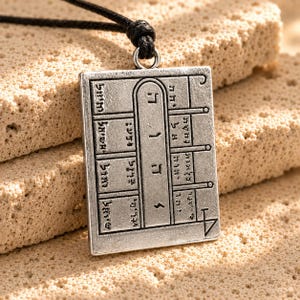 Puede incluir: Un colgante rectangular de metal plateado con un patrón de cuadrícula grabado que contiene varios símbolos y caracteres. El colgante cuelga de un cordón negro y muestra una columna central vertical con caracteres hebreos y líneas.