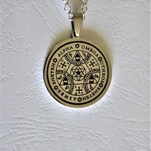 May include: A silver pendant necklace with a round, silver charm. The charm has a black design of a star, a cross, and other symbols. The text "ALPHA" and "OMEGA" are inscribed around the edge of the charm.