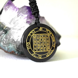 Può includere: Un ciondolo circolare nero con scritte dorate e un design geometrico, sospeso a un cordino nero. Il ciondolo è posto su uno sfondo di cristallo viola e bianco.