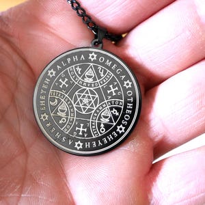 May include: A black metal pendant necklace with a circular design featuring a silver inscription that reads "SEHEYEHASHER TETRA GRAMMATON XOMEGA ALPHA ATON EROEH TETRA GRAMMATON G".