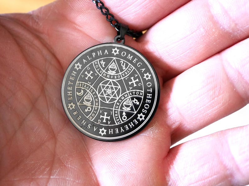 May include: A black metal pendant necklace with a circular design featuring a silver inscription that reads "SEHEYEHASHER TETRA GRAMMATON XOMEGA ALPHA ATON EROEH TETRA GRAMMATON G".