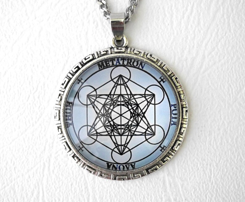 K&ouml;nnte beinhalten: Ein silberfarbener, runder Anh&auml;nger mit einem schwarzen, geometrischen Metatron-W&uuml;rfel-Design auf hellblauem Hintergrund. Der Metallrahmen ist mit einem M&auml;ander-Muster verziert. Der Schriftzug METATRON steht oben, erg&auml;nzt durch weiteren Text.