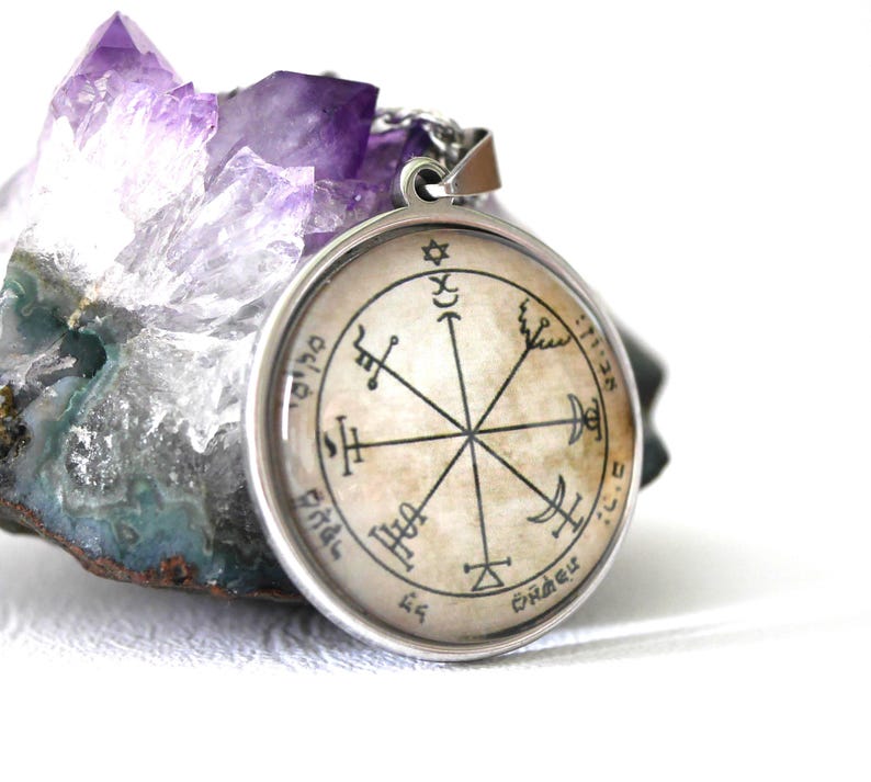 May include: A round pendant with a silver-colored frame and a chain. The pendant features a central design with symbols and text, set against a beige background. The pendant is placed in front of a purple crystal.