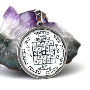 May include: A round, silver-toned pendant with a detailed black and white design, including Hebrew text and symbols. The pendant hangs on a silver chain and rests against a purple crystal. The pendant's design includes the word "Cahetel".