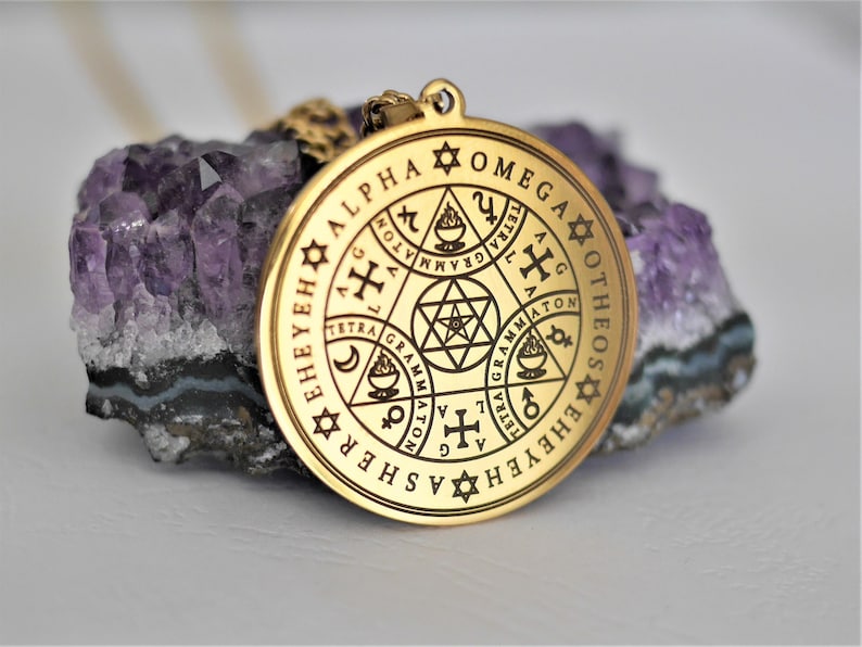 May include: A gold-colored pendant necklace with a circular charm featuring a complex design of symbols and text. The text includes "ALPHA", "OMEGA", "OTHEOS", and "EHEYEH", among others.