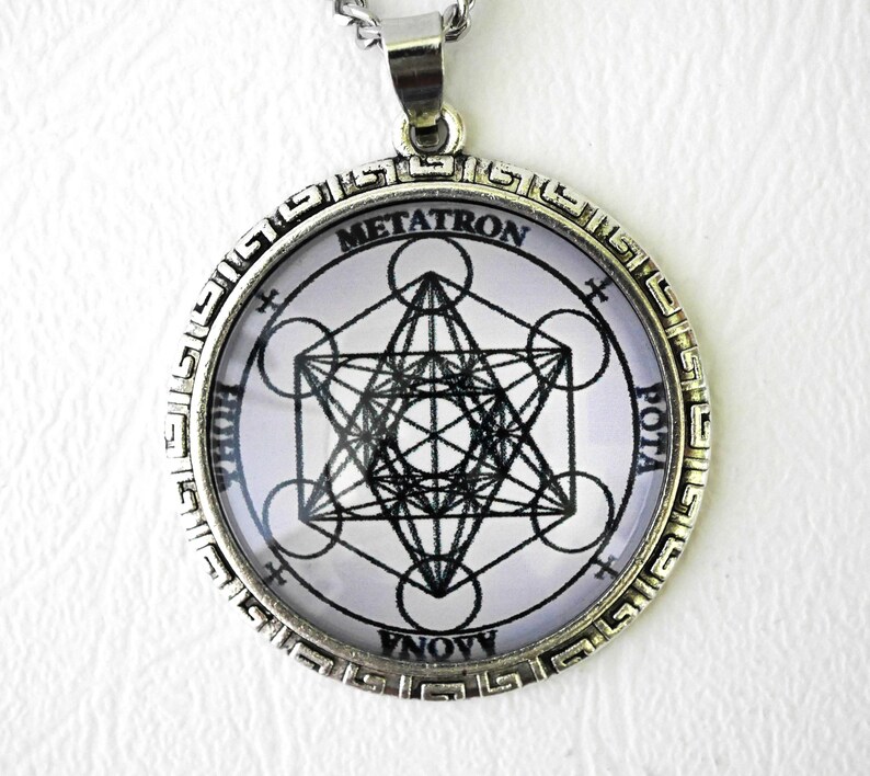 K&ouml;nnte beinhalten: Ein runder, silberfarbener Anh&auml;nger mit einem schwarz-wei&szlig;en geometrischen Metatron-W&uuml;rfel-Design unter einer Glaskuppel. Der Metallrahmen hat einen Rand mit griechischem M&auml;andermuster. Der Text METATRON ist oben aufgedruckt, weitere Buchstaben sind sichtbar.