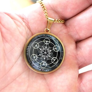 K&ouml;nnte beinhalten: Eine goldfarbene, kreisf&ouml;rmige Anh&auml;ngerkette mit schwarzem Hintergrund und einem wei&szlig;en, geometrischen Metatron-W&uuml;rfel-Design. Der Anh&auml;nger ist in einen glatten Metallrahmen gefasst und h&auml;ngt an einer passenden goldfarbenen Kette. Der Text ADONAY und ROTA ist sichtbar.