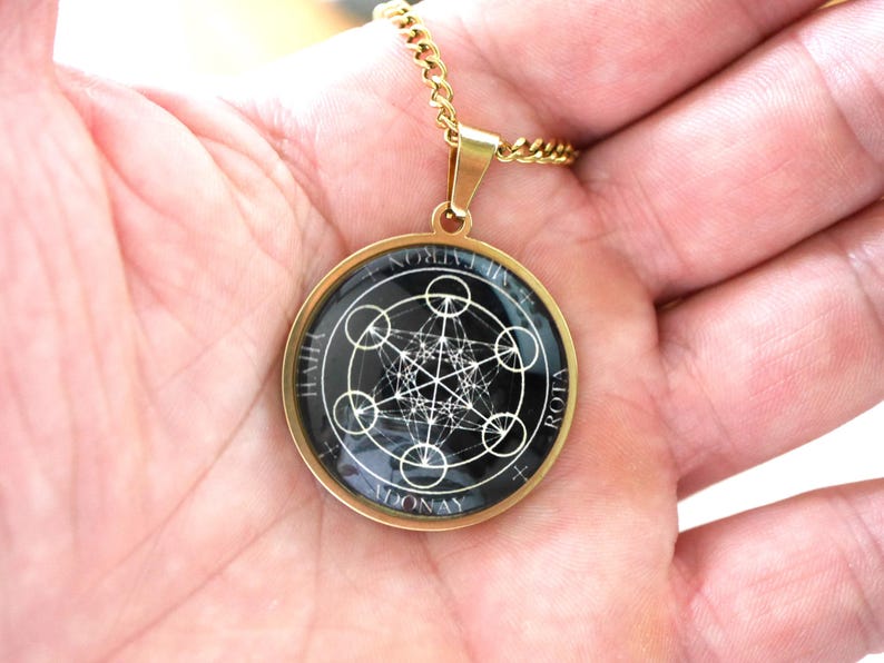 K&ouml;nnte beinhalten: Eine goldfarbene, kreisf&ouml;rmige Anh&auml;ngerkette mit schwarzem Hintergrund und einem wei&szlig;en, geometrischen Metatron-W&uuml;rfel-Design. Der Anh&auml;nger ist in einen glatten Metallrahmen gefasst und h&auml;ngt an einer passenden goldfarbenen Kette. Der Text ADONAY und ROTA ist sichtbar.