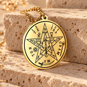 Könnte beinhalten: Ein goldfarbener, runder Anhänger an einer Kette mit einer detaillierten schwarzen Gravur eines Pentagramms. Der Anhänger zeigt verschiedene Symbole wie ein Schwert, einen Kelch sowie den Text "1 2 3 TRA" und "GRAM MATON" am Rand.