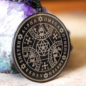 May include: A black metal pendant necklace with a circular design featuring a star of David, a cross, and the words "Alpha Omega Otheos Ehyehashe" inscribed around the edge.