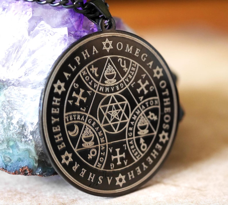 May include: A black metal pendant necklace with a circular design featuring a star of David, a cross, and the words "Alpha Omega Otheos Ehyehashe" inscribed around the edge.