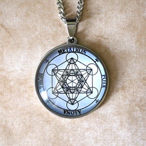 K&ouml;nnte beinhalten: Ein runder silberfarbener Anh&auml;nger mit einer schwarz-wei&szlig;en geometrischen Metatron-W&uuml;rfel-Grafik unter einer Glaskuppel. Der kreisf&ouml;rmige Rahmen enth&auml;lt die Schriftz&uuml;ge METATRUN, HUHA, PUIA und AONA um das zentrale geometrische Symbol.