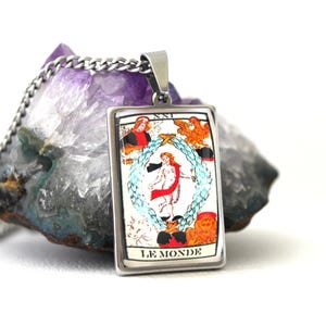 May include: A silver-toned pendant necklace featuring a rectangular charm with a tarot card design. The card depicts a figure within a laurel wreath, with the words "LE MONDE" at the bottom. The pendant hangs on a silver chain, set against a purple and gray stone.