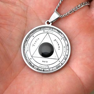 Può includere: Una collana d'argento con un ciondolo rotondo che presenta una pietra nera al centro di un triangolo. Il ciondolo ha una scritta ebraica attorno al bordo, tra cui le parole "Raphael", "Michael", "Auriel", "Tetra" e "Gramma".