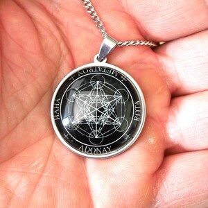 K&ouml;nnte beinhalten: Ein runder silberfarbener Anh&auml;nger mit einem schwarzen kreisf&ouml;rmigen Einsatz, der ein wei&szlig;es geometrisches Metatron-W&uuml;rfel-Design zeigt. Der Rand des Anh&auml;ngers ist mit dem Text METATRON, YHVH, ADONAY und BOTA in wei&szlig;en Gro&szlig;buchstaben versehen.