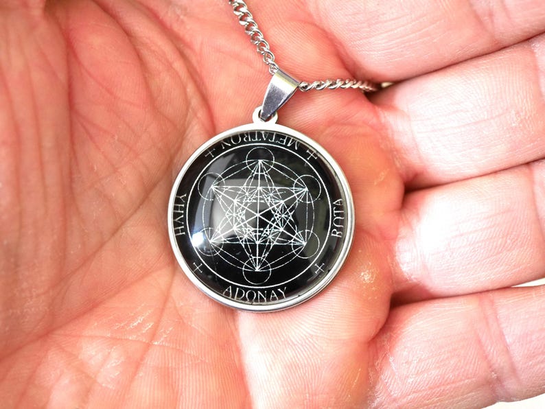 K&ouml;nnte beinhalten: Ein runder silberfarbener Anh&auml;nger mit einem schwarzen kreisf&ouml;rmigen Einsatz, der ein wei&szlig;es geometrisches Metatron-W&uuml;rfel-Design zeigt. Der Rand des Anh&auml;ngers ist mit dem Text METATRON, YHVH, ADONAY und BOTA in wei&szlig;en Gro&szlig;buchstaben versehen.