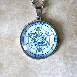 K&ouml;nnte beinhalten: Ein kreisf&ouml;rmiger silberfarbener Anh&auml;nger mit einem blauen geometrischen Metatron-W&uuml;rfel-Design auf wei&szlig;em Hintergrund. Der Text METATRON ist oben aufgedruckt, mit zus&auml;tzlichem Text entlang des Umfangs des kreisf&ouml;rmigen Rahmens.