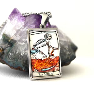 Peut inclure: Pendentif rectangulaire en argent avec un motif de carte de tarot. La carte représente un squelette avec une faux, sur fond orange et rouge. Le texte "LA MORT" est en bas. Le pendentif est suspendu à une chaîne en argent.