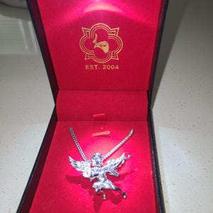 May include: A silver-colored pendant necklace featuring an angel with wings and a weapon, displayed in a red velvet-lined box. The box is black with gold lettering and the text "EST. 2004".