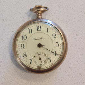 Op de afbeelding: Een goudkleurig Hamilton zakhorloge met een crèmekleurige wijzerplaat. Het horloge heeft zwarte cijfers, een kleine secondenwijzer en de merknaam "Hamilton" in script. Het heeft een kroon aan de bovenkant en een secondewijzer.