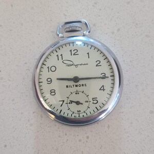 May include: A silver-toned Ingraham Biltmore pocket watch with a cream-colored face. The watch features black numerals, hands, and a subdial. The watch face displays the brand name and the words "USA".