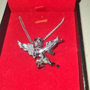 May include: Silver-toned pendant necklace featuring a cherub with wings, holding a rifle. The pendant is displayed in a red jewelry box. The chain is a delicate link style.