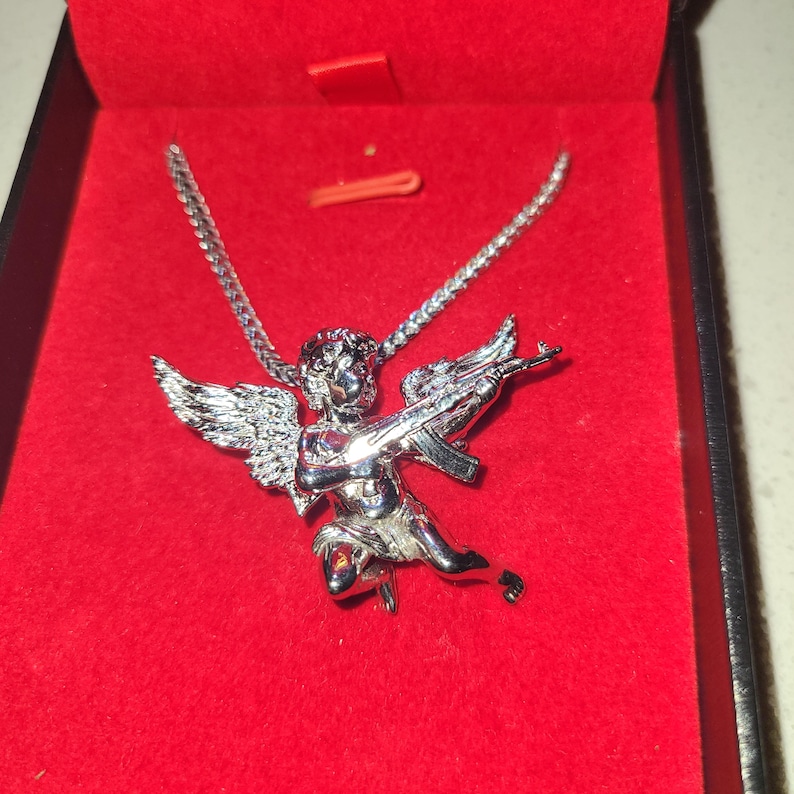 May include: Silver-toned pendant necklace featuring a cherub with wings, holding a rifle. The pendant is displayed in a red jewelry box. The chain is a delicate link style.