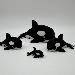 May include: A collection of black and white orca plush toys in various sizes. The orcas have white bellies and eye patches, with a smiling expression. The toys are arranged on a white surface.